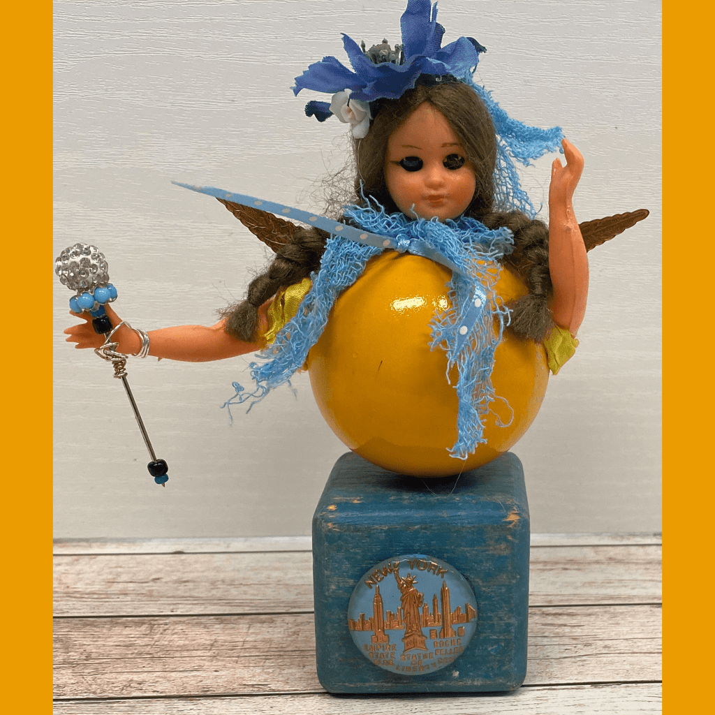 A whimsical doll dressed in yellow, standing on a blue box labeled 'NEW YORK'. The doll has brown hair adorned with flowers and wears a blue scarf. She holds a shiny wand in one hand and has orange wings made of ice cream cones.