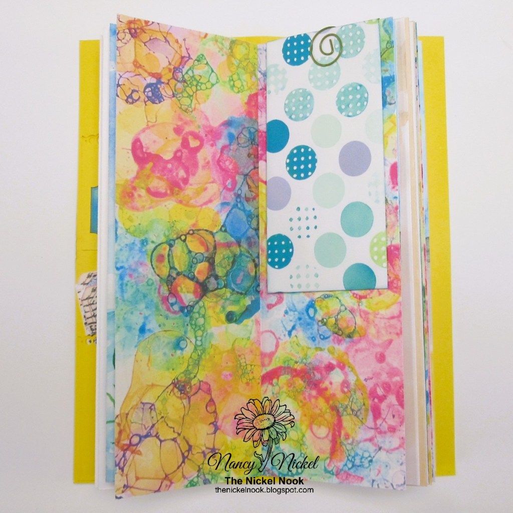 An open junk journal page with a colorful background featuring bright, abstract patterns in pastel colors, including shades of teal, pink, and yellow. A decorative tag with blue and green circular designs is tucked into the side.