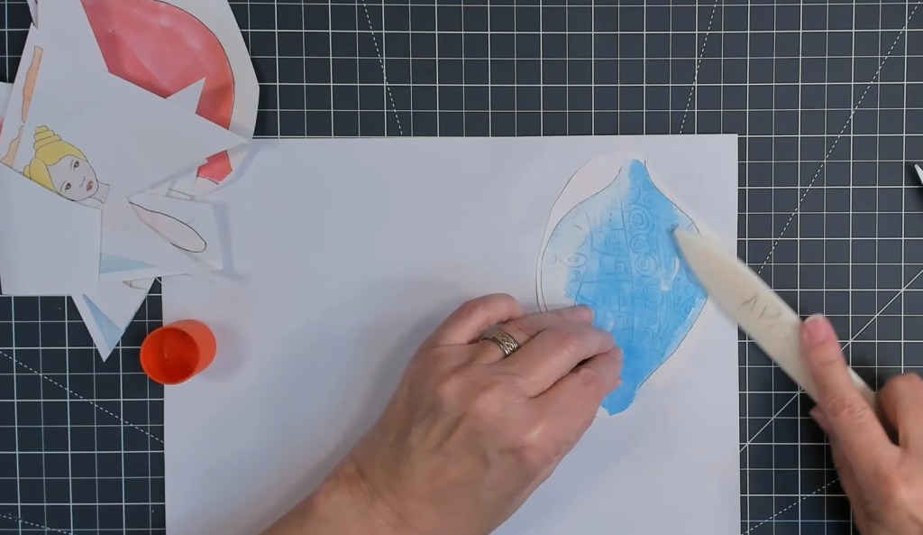 A person's hand applying glue to a colorful, mermaid-themed paper cutout on a crafting workspace, with various mermaid designs in the background.