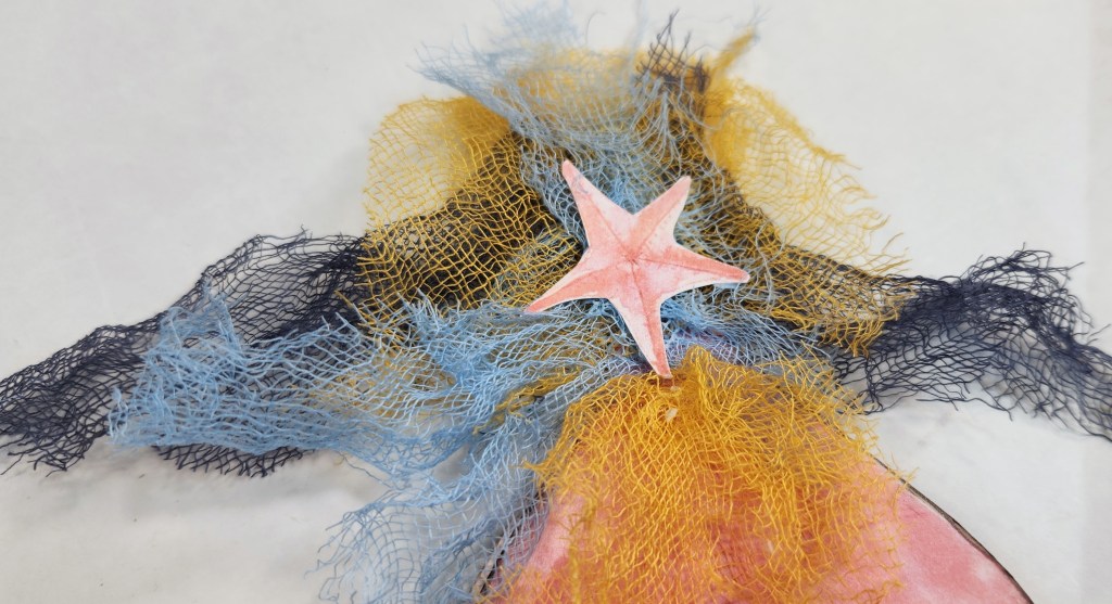 A close-up view of a colorful craft piece featuring layers of colored cheese cloth in blue, yellow, and black, with a pink starfish element on top.
