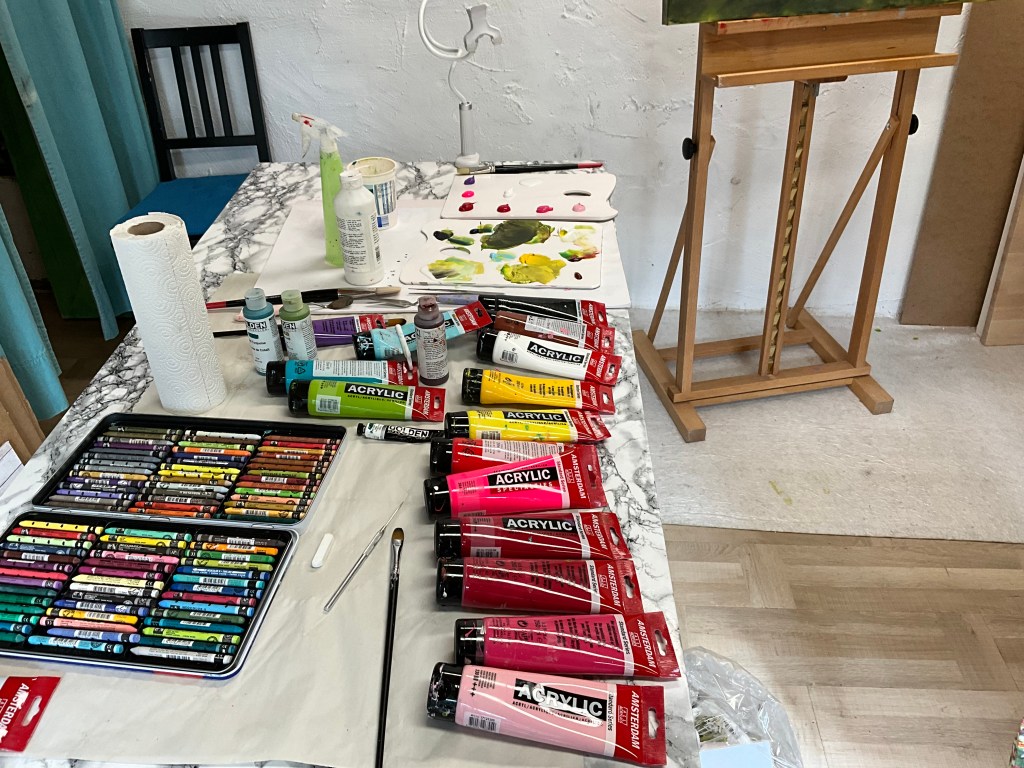 Art studio table setup with various acrylic paints, Neopastel crayons, brushes, and mixing palette used for the mixed media painting Rosefields