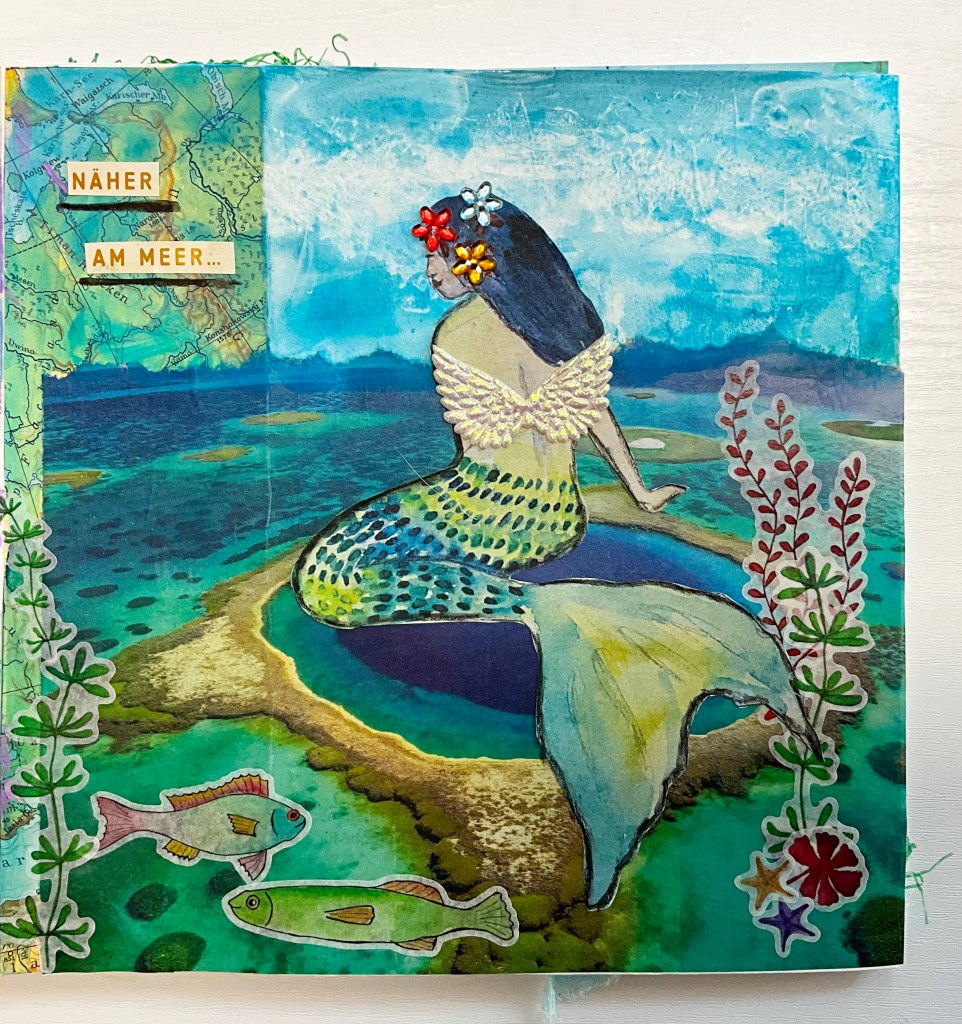 A vibrant ocean-inspired art journal page featuring a mermaid with a multicolored tail sitting by the water. The background includes a mix of a torn map and a bright blue sky. Decorative washi stickers of sea life and a quote 'Näher am Meer...' are included.