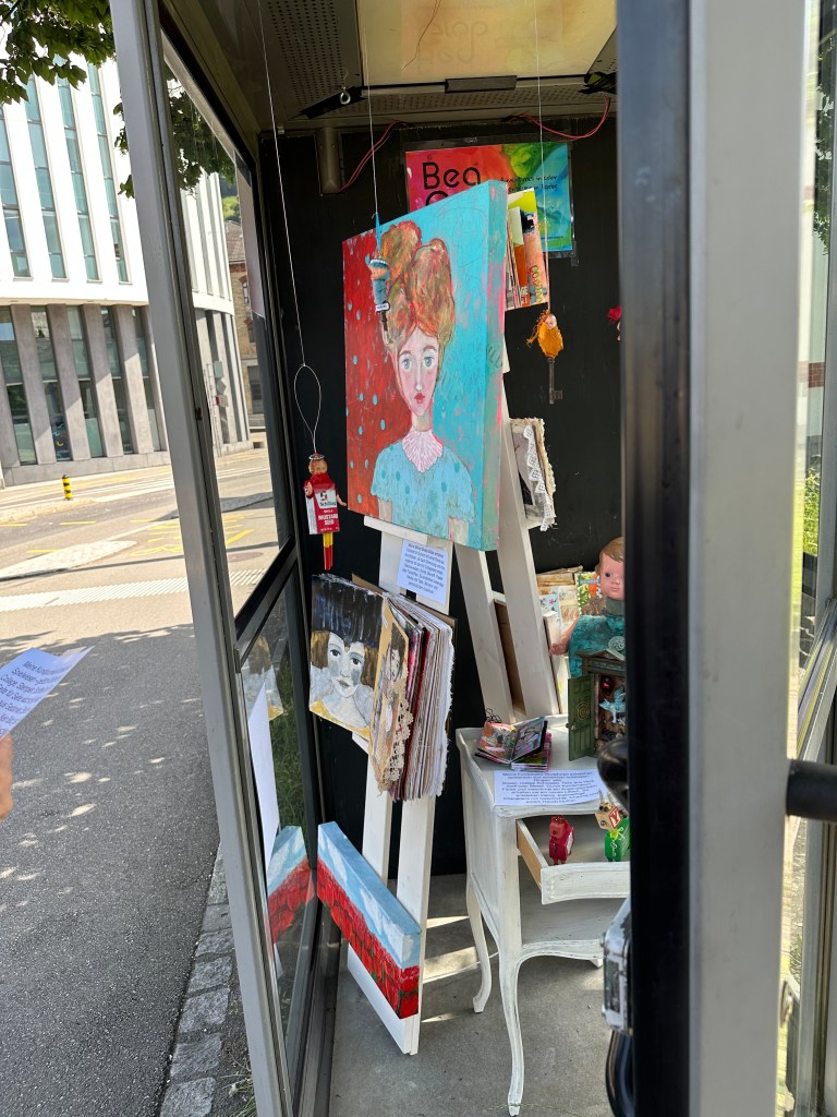 View inside the phone booth exhibition showing paintings, art journals, and a small doll assemblage.nterior view of a public phone booth mini-exhibition featuring artworks, including paintings, art journals, and small assemblages. The setup includes a prominently displayed colorful portrait and various art pieces on stands.
