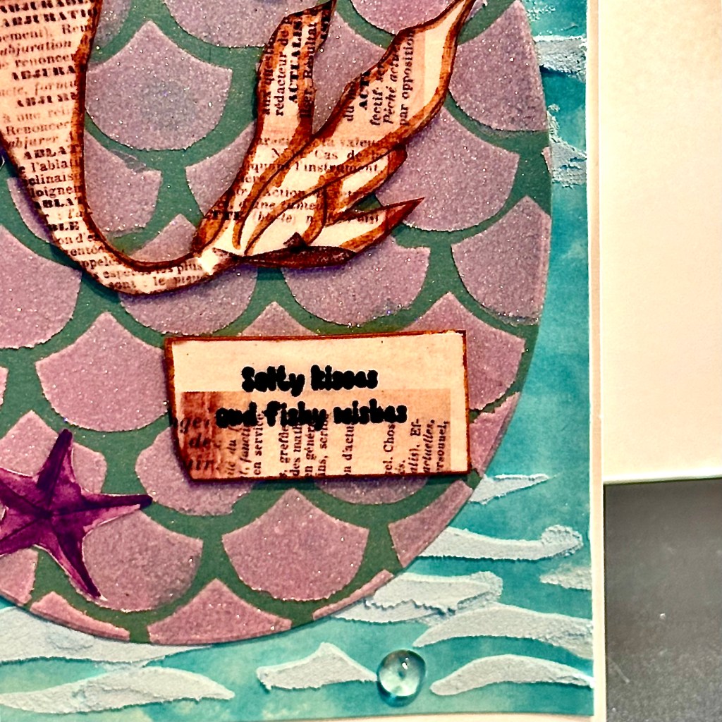 A layered card featuring a mermaid tail made from vintage-style paper, set against a shimmering purple-scaled background and adorned with sea-themed ephemera and the sentiment 'Salty kisses and fairy wishes'.