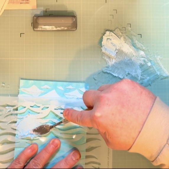 A person applies texture paste to a stencil depicting ocean waves, using a palette knife on a crafting surface.