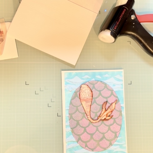 A colorful card featuring a shimmering purple-scaled backdrop and a vintage-style mermaid tail in the center, surrounded by ocean wave stenciling and playful ephemera elements.