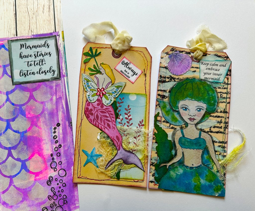 A whimsical art journal spread featuring two handmade tags with mermaid illustrations, decorative elements like butterflies and seashells, and a colorful background with various textures.