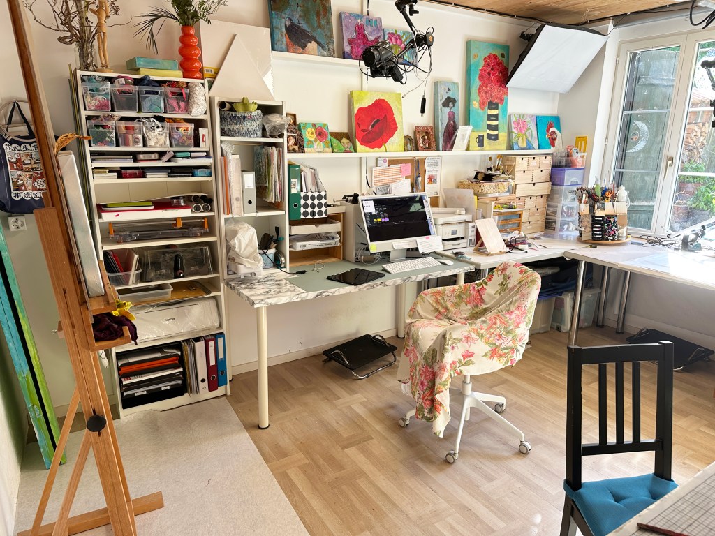 A bright and organized art studio featuring a desk with a computer, colorful artwork on the walls, shelves filled with crafting materials, and a comfortable chair draped with a floral fabric.