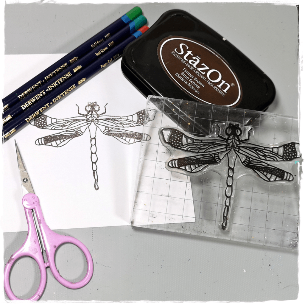 A workspace showcasing a dragonfly stamp on a clear block next to a drawing of a dragonfly, colored pencils, scissors, and StazOn ink pad.