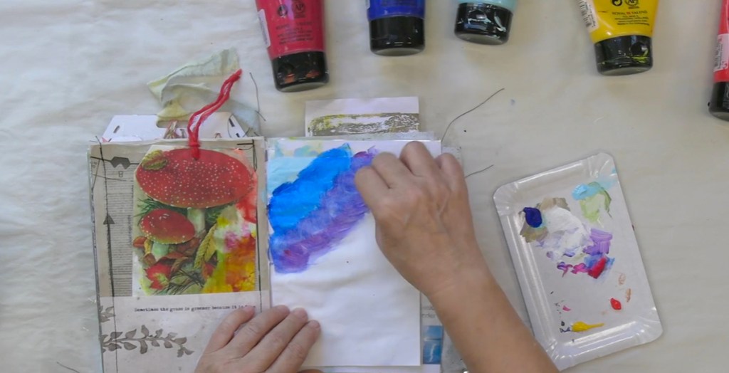 Eine Hand zeigt Farben auf einer offenen Mixed-Media-Seite, umgeben von verschiedenen Acrylfarben in Tuben und einer Farbpalette.