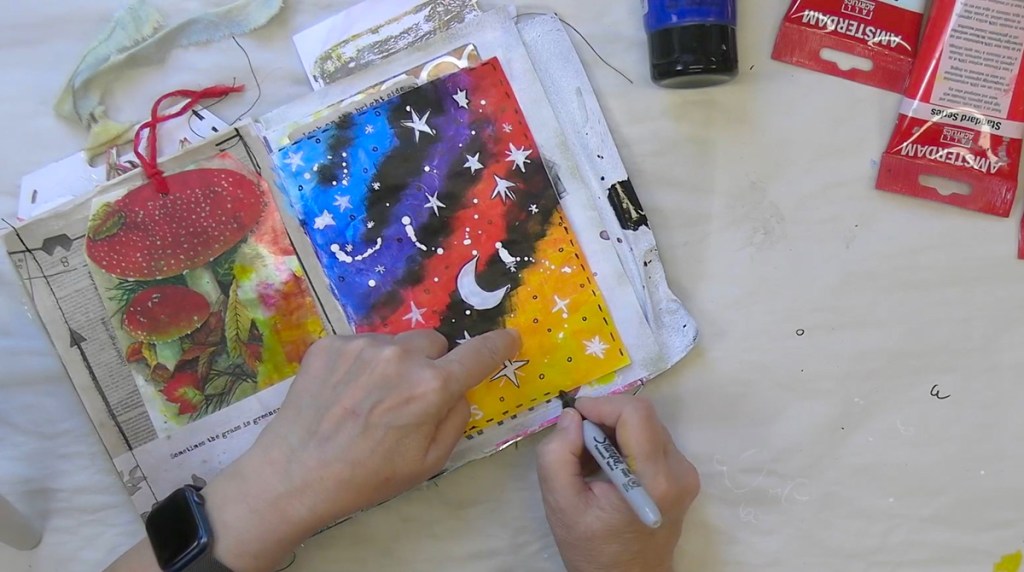 Ein kreativer Kunstprozess, bei dem eine Person mit Acrylfarben und einem Marker eine bunte, galaktische Hintergrundlein auf einer durchsichtigen Seite eines Notizbuchs gestaltet.