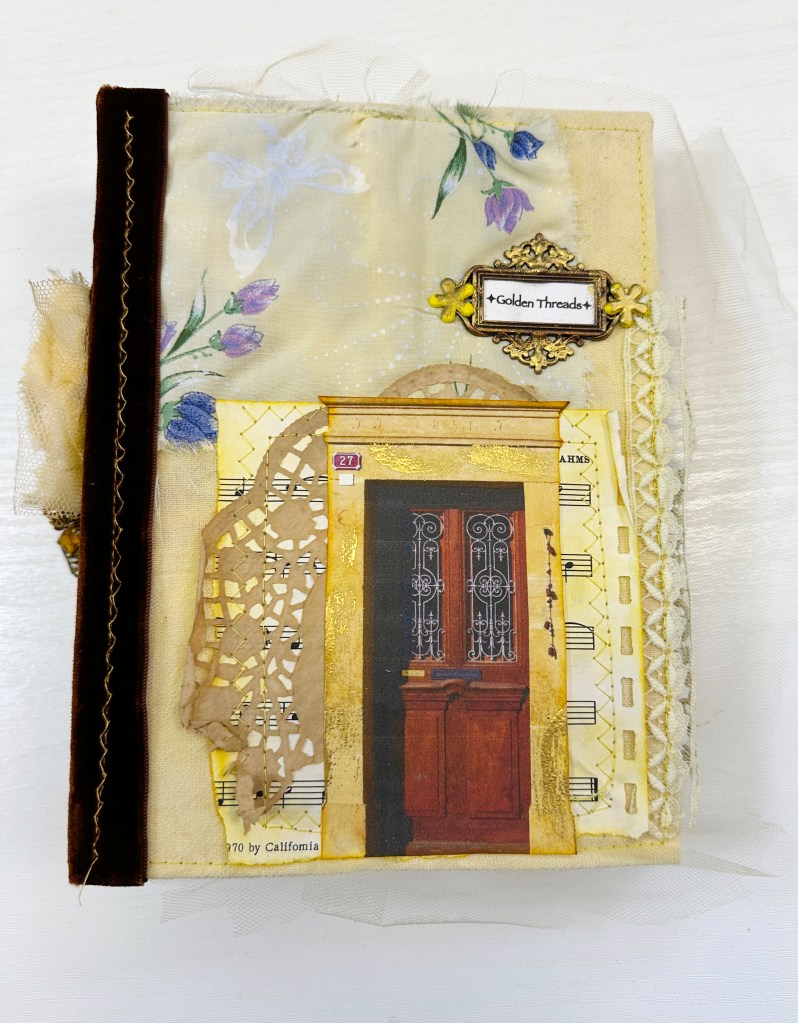 A close-up of a handmade art journal cover featuring a decorative design with flowers, lace, and a prominent door image. The journal is adorned with a label reading 'Golden Threads' and various textures including fabric and paper.