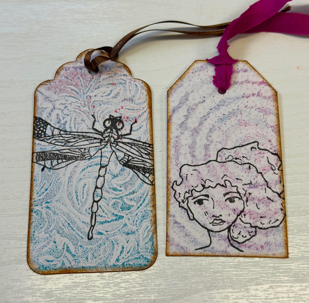 Two decorative tags featuring artwork: one with a black and white illustration of a dragonfly on a colorful background, and the other with a sketched face of a woman. Each tag has a textured design and is finished with a ribbon.