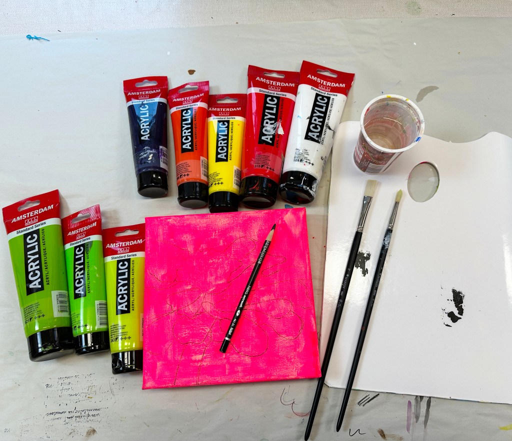 Art supplies arranged on a table, including various acrylic paint tubes, brushes, a cup of water, and a pink canvas with a drawn pattern.