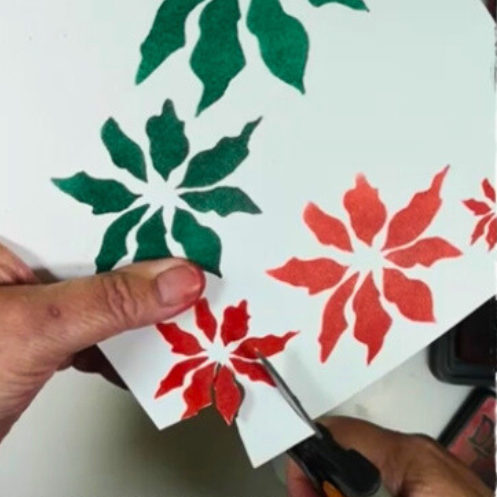 A close-up image of a person cutting out handmade Poinsettia flowers from a stencil, featuring red and green petals against a white background.