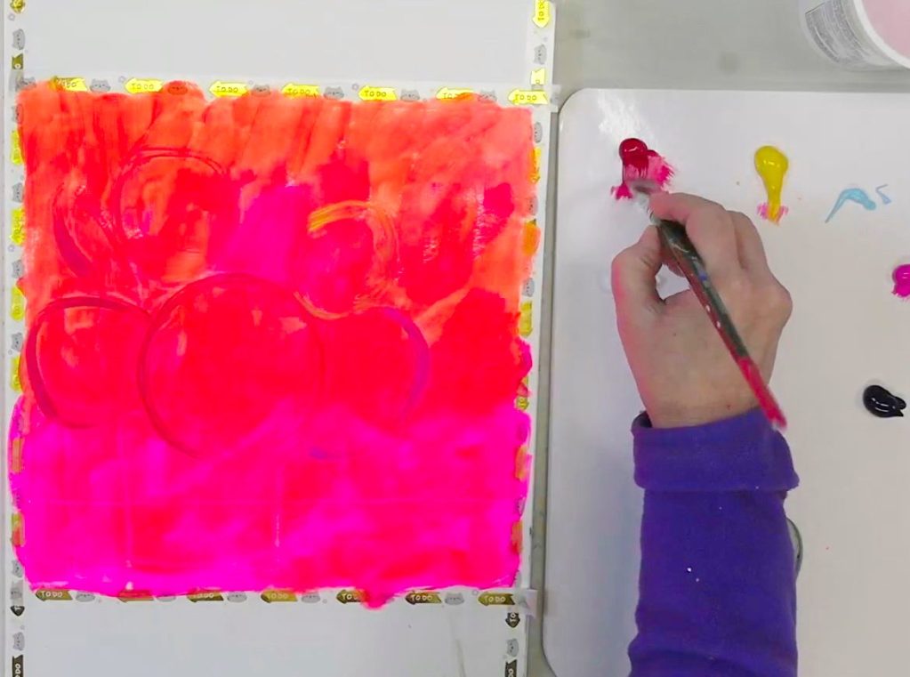 An artist's hand painting a vibrant fluorescent pink and orange background on A3 watercolor paper in a Moleskine Art Journal.