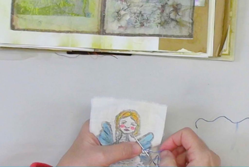 A person hand-stitching a small fabric piece featuring a winter fairy, with a journal open in the background showcasing textured collage pages.