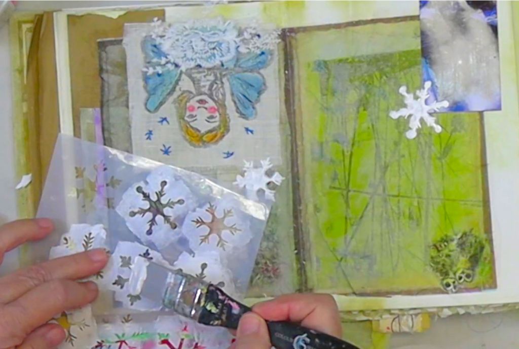 An artist's hands applying paint using a snowflake stencil over a mixed media art page featuring a hand-drawn winter fairy and layered textures.