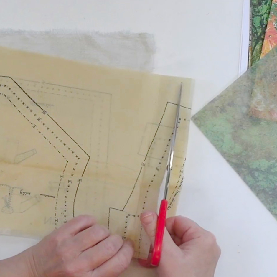 A person's hands using scissors to cut a sewing pattern from a translucent sheet of paper, with fabric and other colorful papers visible in the background.
