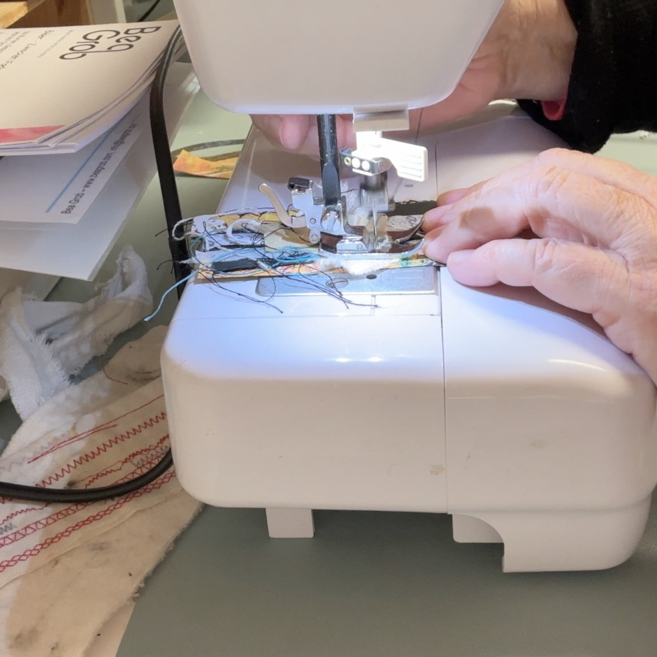 A person's hand guiding fabric through a sewing machine with threads and fabric scraps visible.