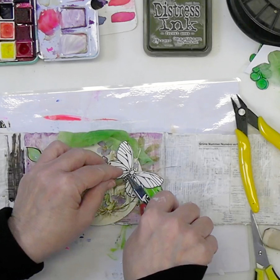 A person's hands cutting out a butterfly design from paper, surrounded by art supplies including watercolor paints, ink, and scissors on a work surface.