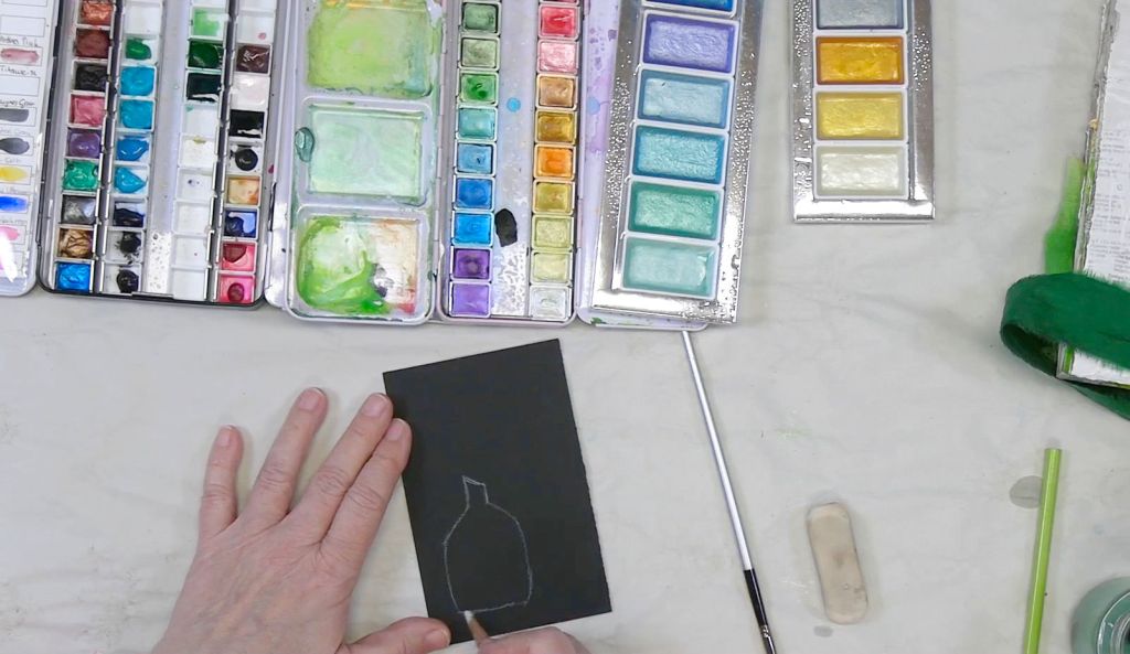 An artist's hand sketching a vase outline on black paper, surrounded by various watercolor palettes and brushes.