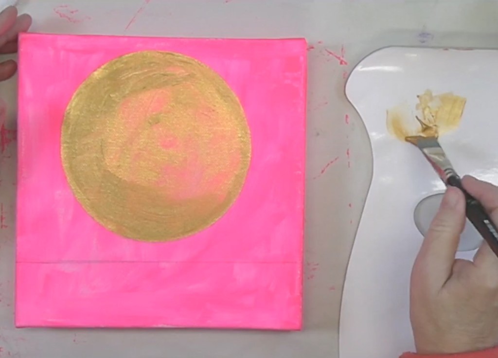 An artist's hand holding a paintbrush over a palette with a golden paint, working on a canvas featuring a bright pink background and a large gold circle.