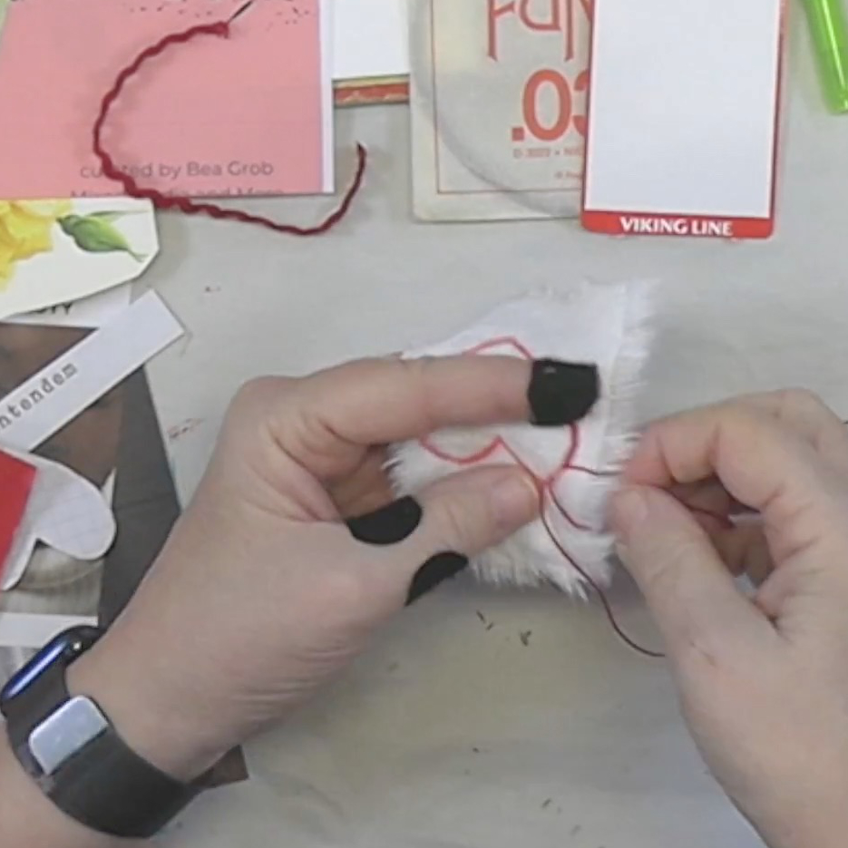 A close-up of hands sewing red thread onto a piece of white fabric, surrounded by crafting materials and colorful papers.