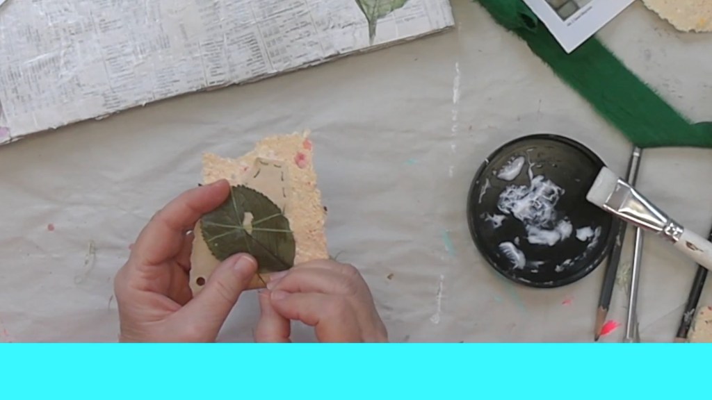 A person holds a piece of handmade paper with a green leaf glued on it, surrounded by crafting materials including a paintbrush and a circular container with white paint.