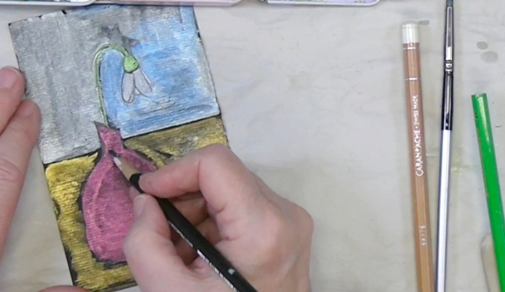 A person's hand coloring a small artwork featuring a pink vase and a snowdrop flower against a blue background, with colored pencils and brushes nearby.