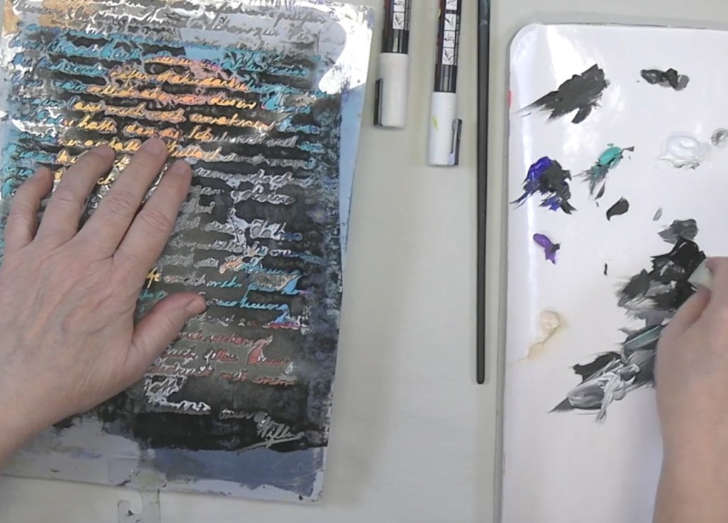 A close-up view of a person's hands working on an art project, one hand resting on a textured surface with handwritten notes, while the other hand holds a paintbrush near a palette filled with various paint colors.