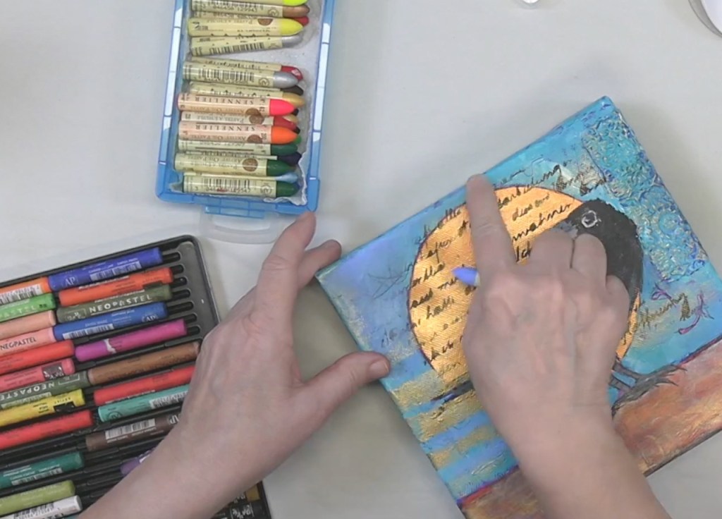 An artist's hand using a blue marker to draw on a colorful canvas featuring a bird and a large yellow sun, with several art supplies in the background.
