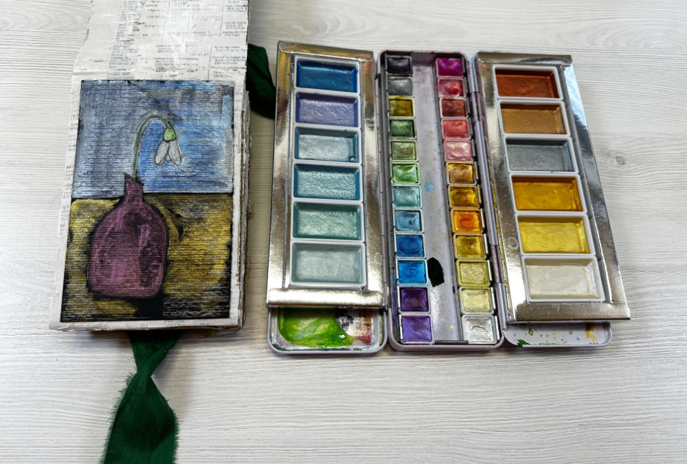 An open sketchbook featuring a colorful drawing of a snowdrop flower in a purple vase on a textured background, accompanied by two palettes of watercolor paints with a variety of vibrant colors.