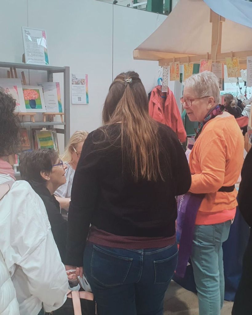 A group of people engaged in conversation at a trade show or craft fair, with one elderly woman wearing glasses and an orange top talking to attendees while others are seated and listening.