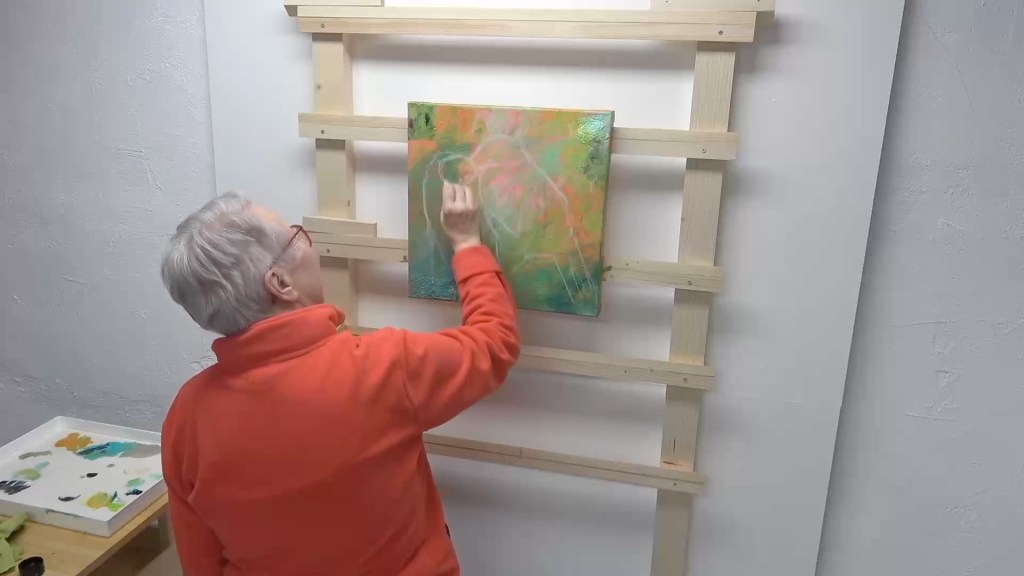 An elderly artist in an orange sweater is painting on a colorful canvas mounted on a wooden easel in a well-lit studio.