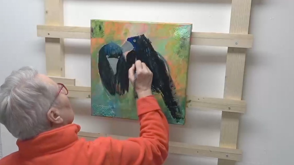 An artist painting a vibrant canvas featuring two black birds, with a colorful abstract background, while seated and wearing an orange sweater.
