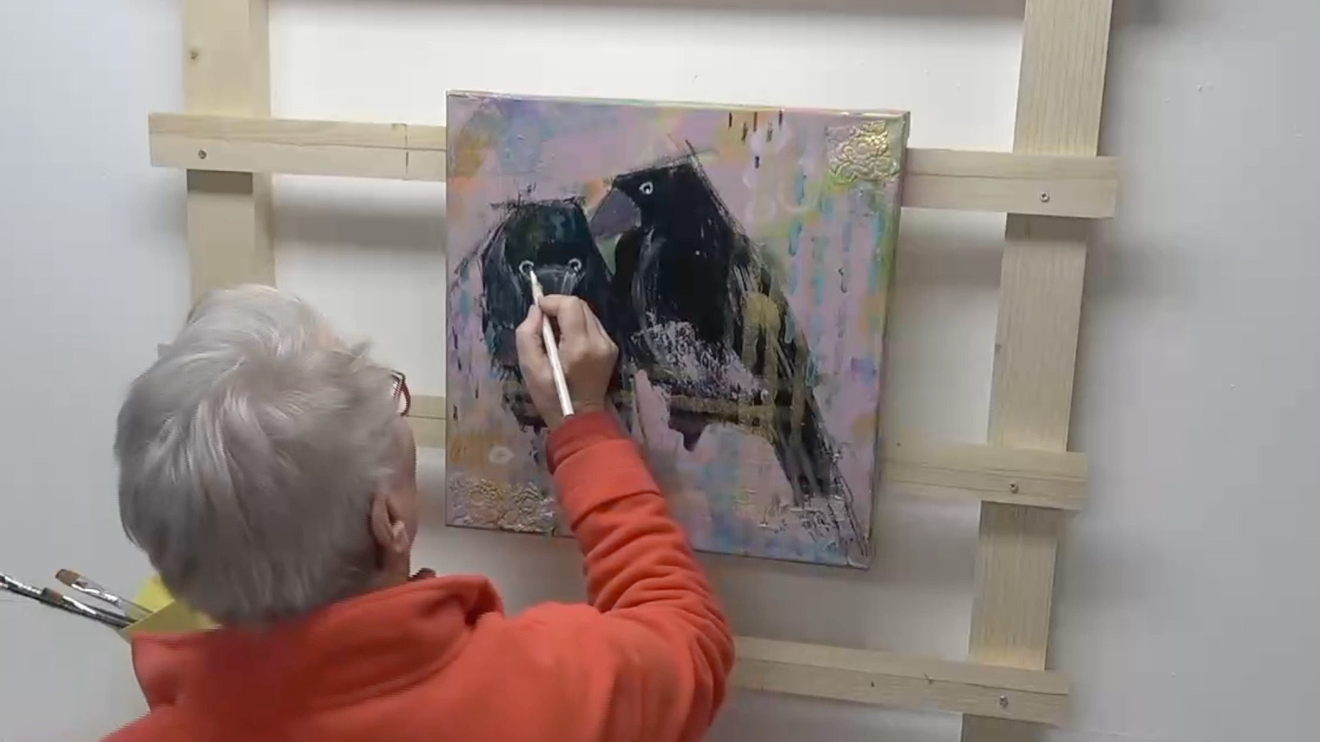 An artist painting a colorful artwork featuring two birds on a canvas mounted to a wooden easel.