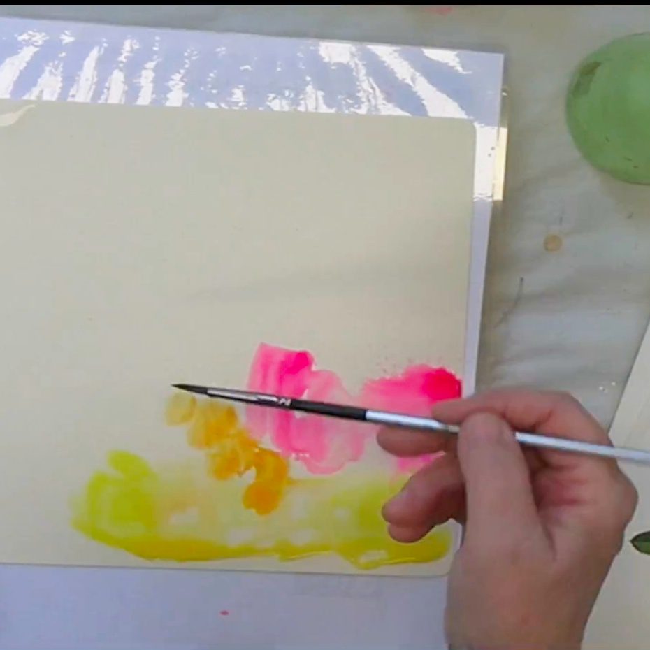 A person holding a paintbrush, applying vibrant colors such as pink, yellow, and orange onto a blank canvas. The workspace includes a light-colored background and hints of other art materials.