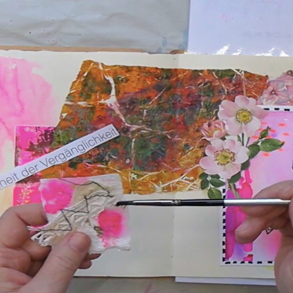 A close-up of a hand holding a small piece of fabric with pink and beige colors while painting with a brush. Brightly colored collage elements like floral designs and handwritten text are visible in the background on a sketchbook page.