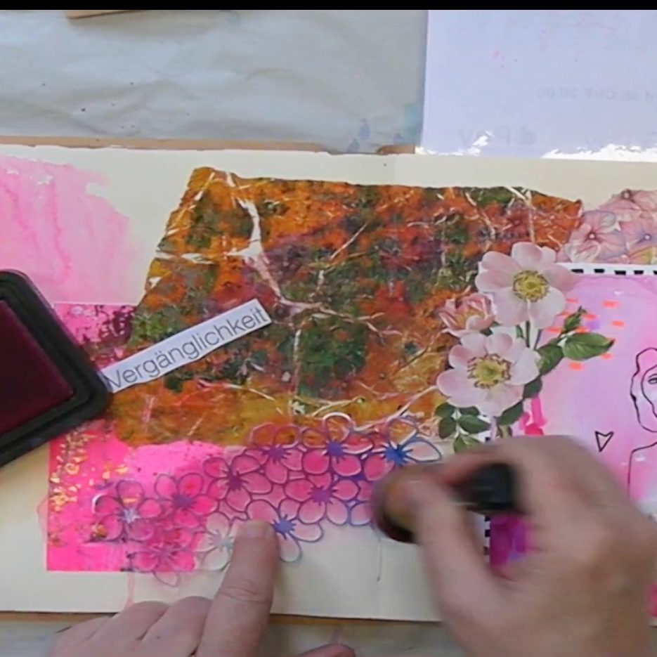 A close-up of a mixed media art journal page featuring colorful floral designs, overlapping textures, and the word 'Vergänglichkeit' in German. Hands apply ink with a stamp near the bottom of the page.
