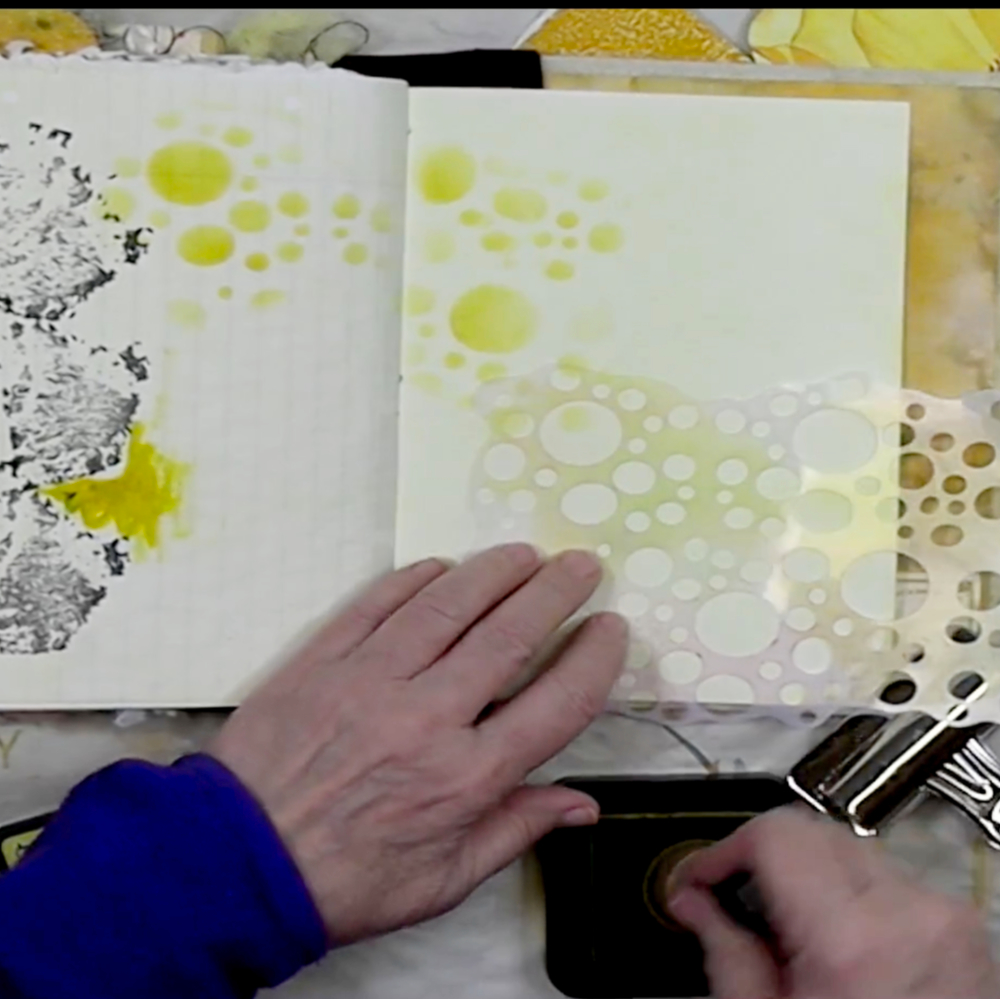 A person's hand applying a stencil with bubble patterns on a blank page in a notebook, surrounded by yellow ink splatters.