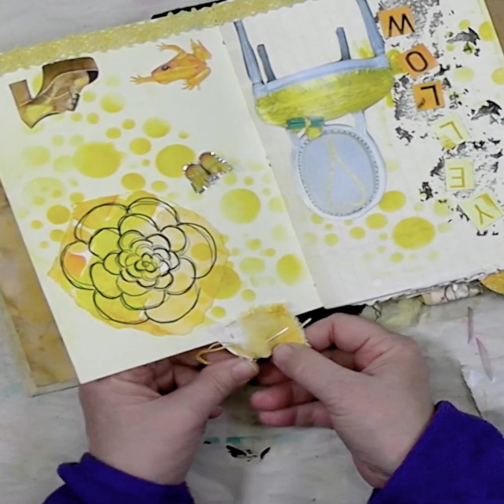An open art journal displaying yellow-themed pages with a flower illustration, a cartoon frog, and the word 'Yellow' in colorful letters. A hand holds a piece of fabric with thread.