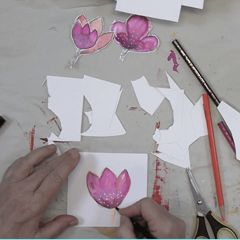 A person's hand coloring a pink flower illustration on a piece of white paper, surrounded by cut-out flower shapes and art supplies like scissors and colored pencils.