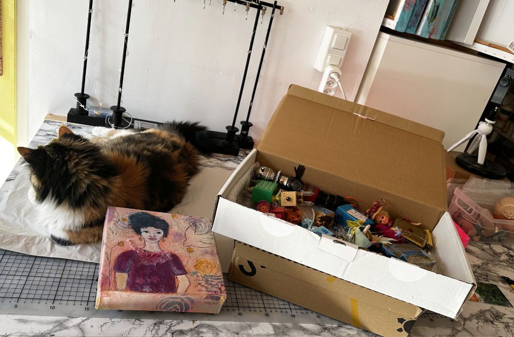 A calico cat resting on a marble surface next to a colorful box and an open cardboard box filled with various small items and toys.