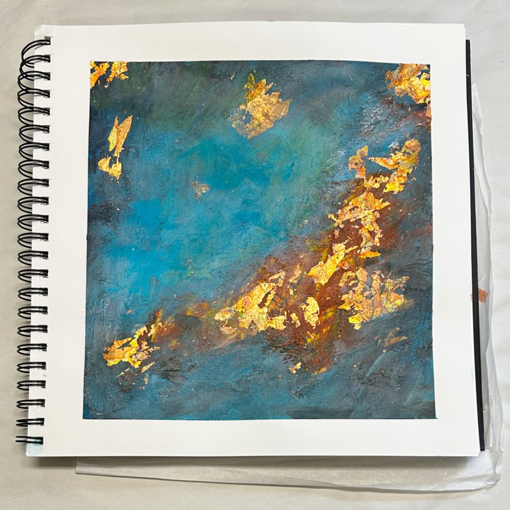 An abstract painting featuring a blend of blue, turquoise, and brown colors, embellished with gold leaf accents. The artwork is presented on a white paper background in a spiral-bound sketchbook.