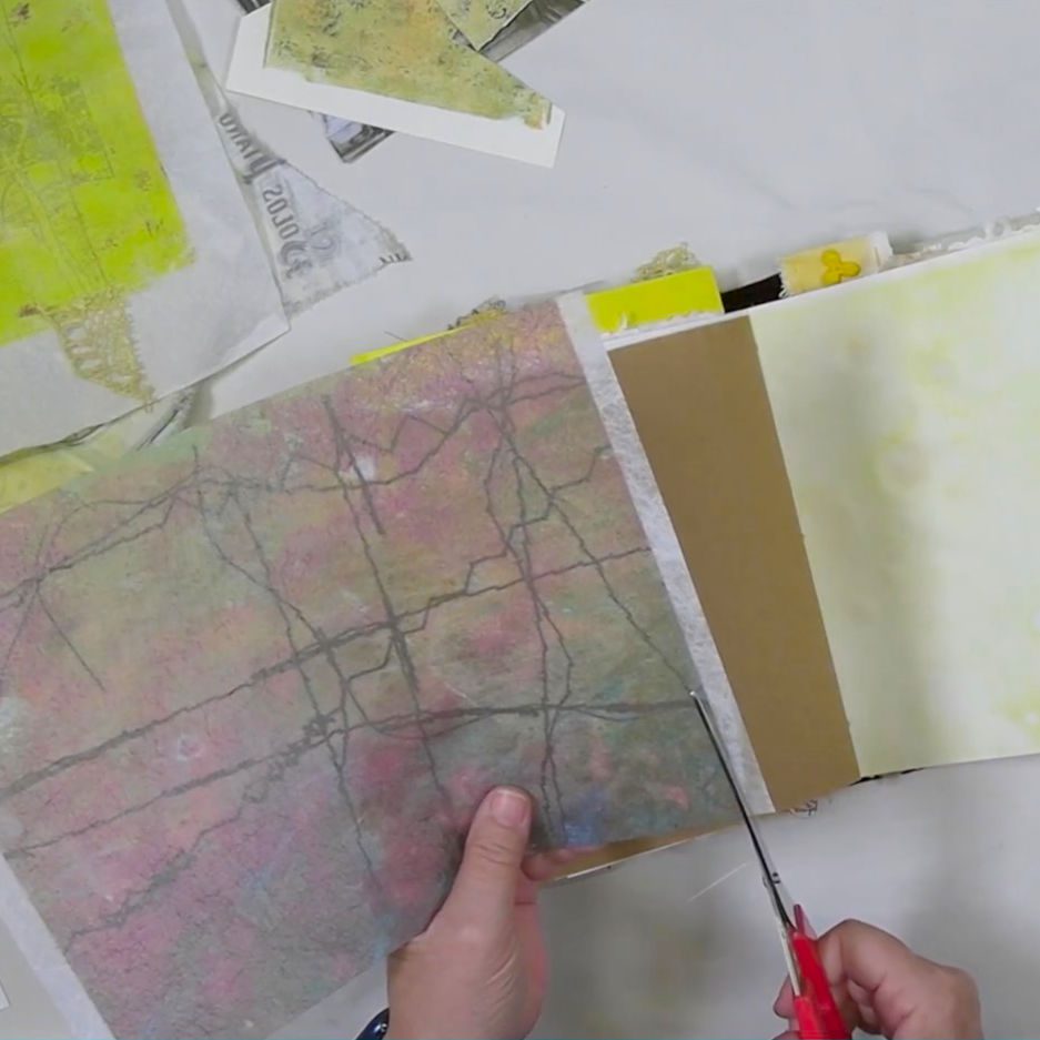 Person holding a sheet of colorful, patterned paper while cutting it with scissors, surrounded by various other crafting materials on a table.