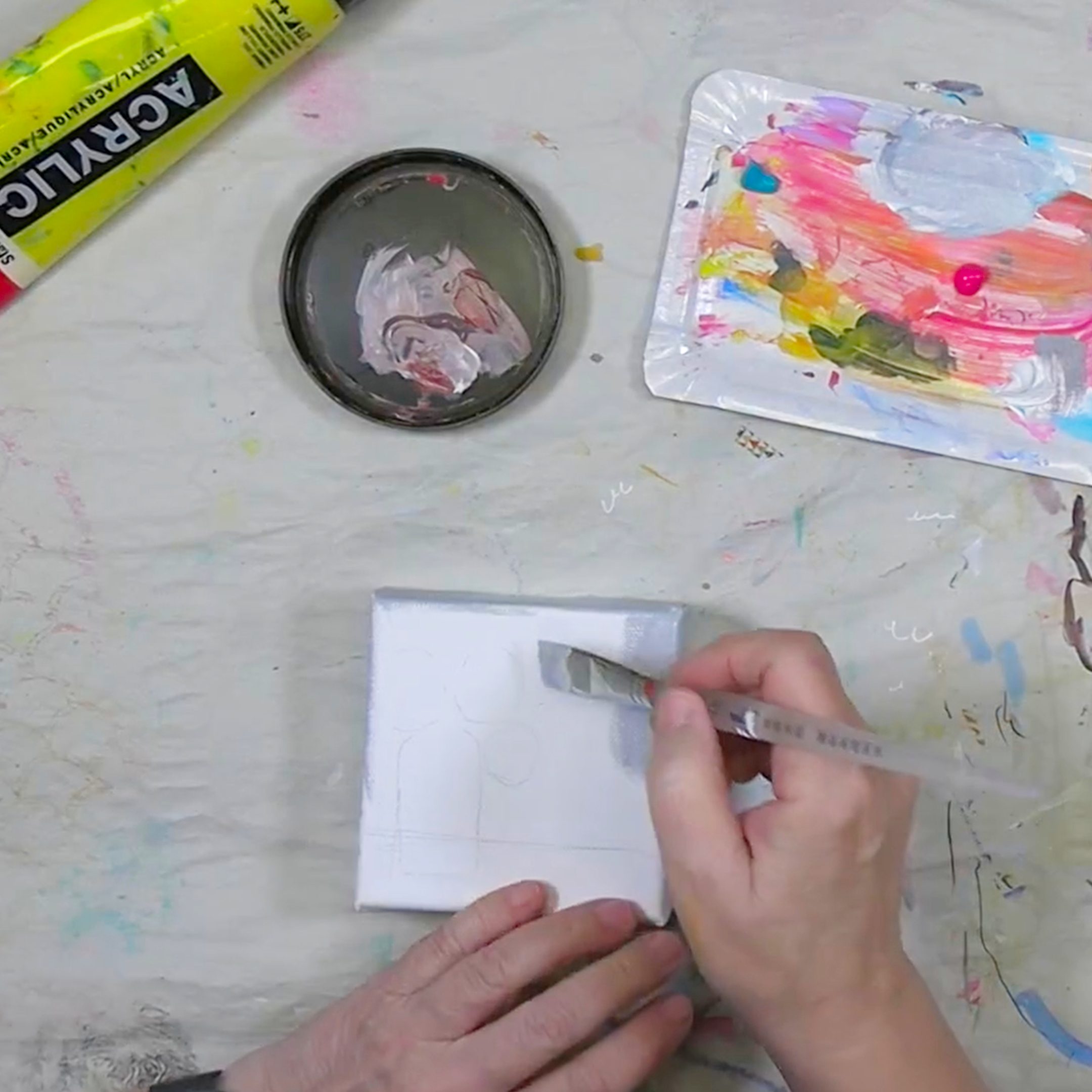An artist's hand applying paint to a small canvas with a palette and paint tubes visible in the background.