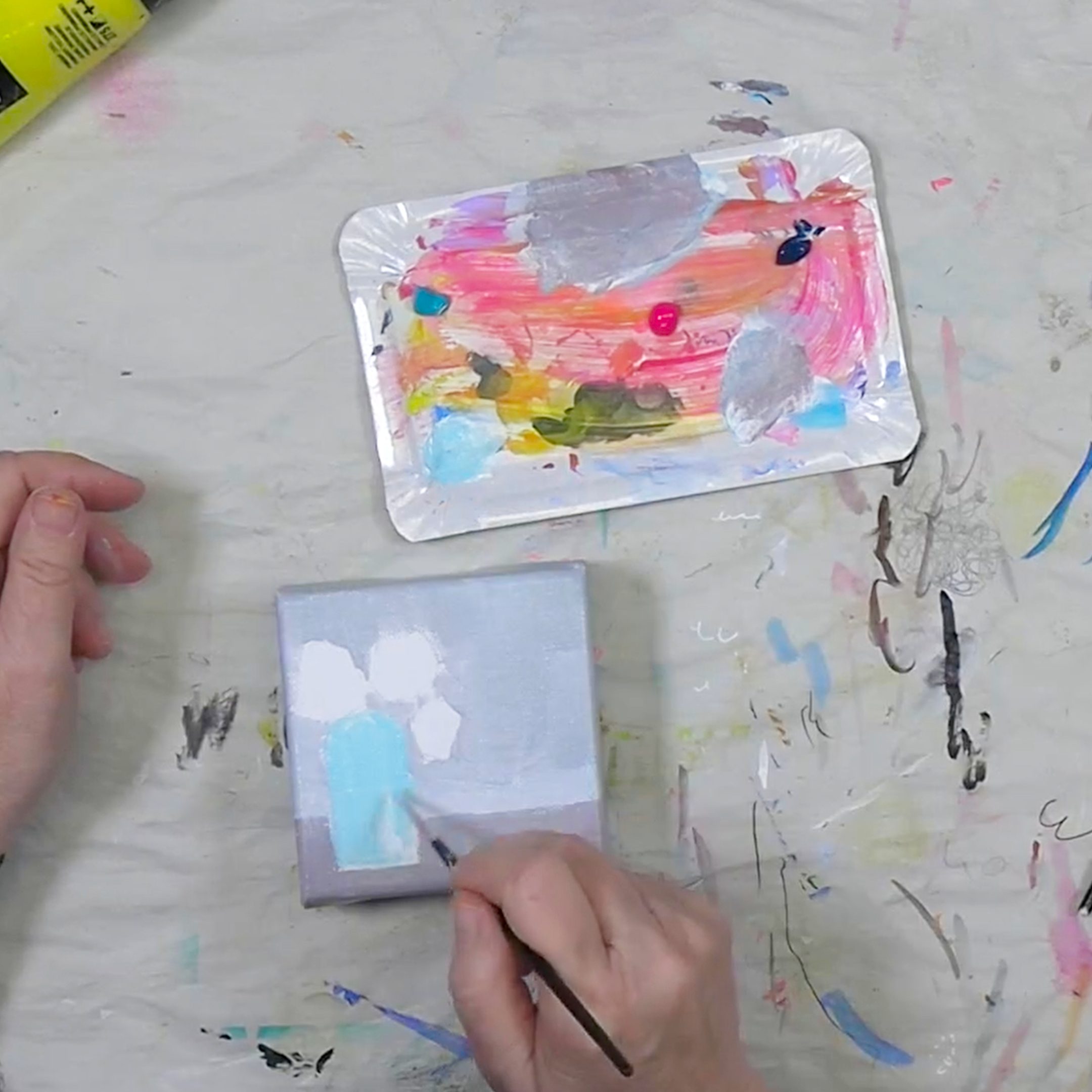 An artist's hand holding a paintbrush, working on a small canvas with light blue and white paint, alongside a paint palette with various vibrant colors.