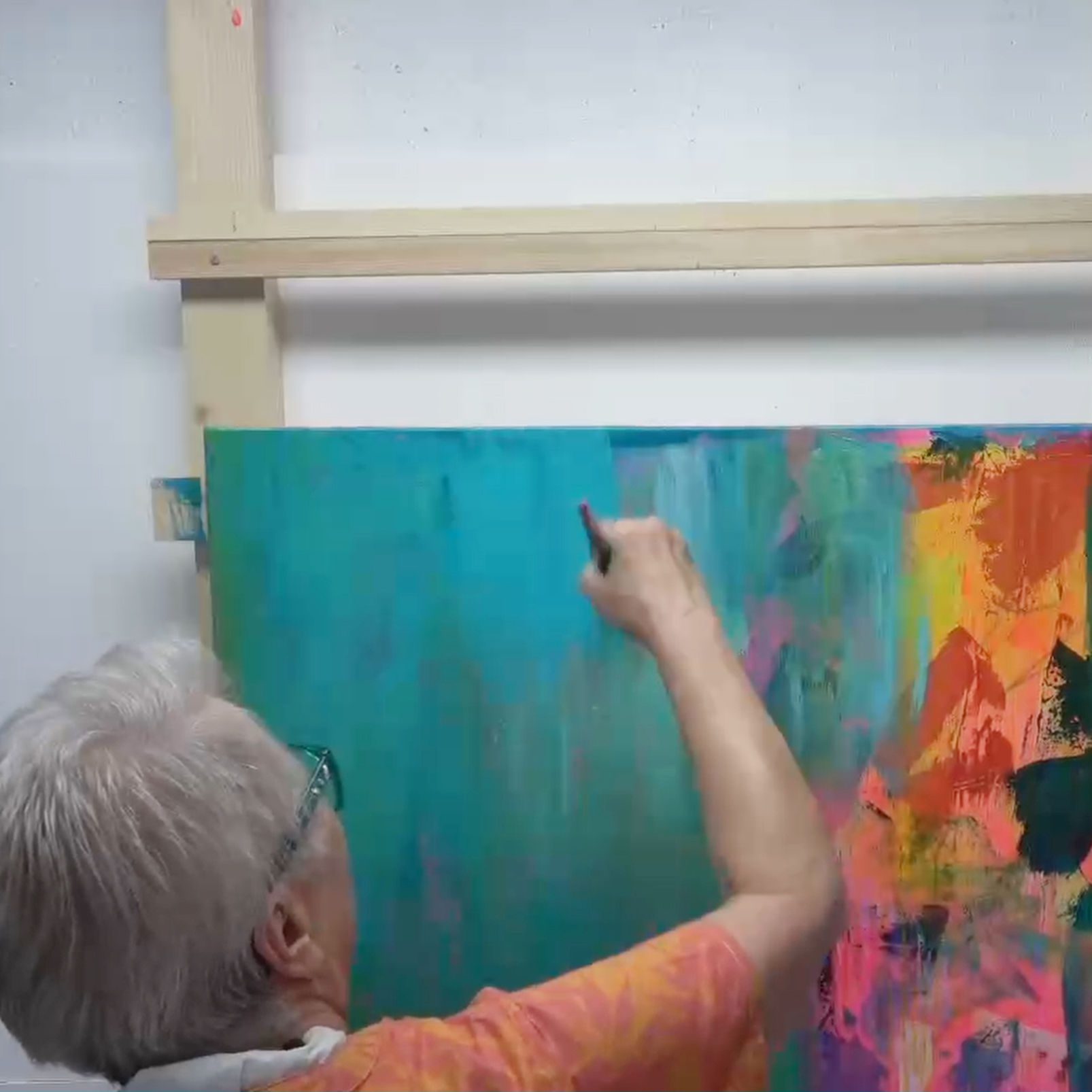 An artist painting on a large canvas with vibrant colors, focusing on a turquoise section, wearing an orange shirt and glasses.