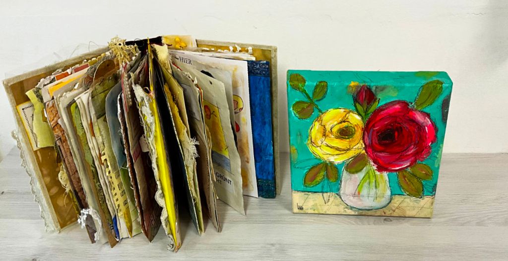An open artists' book filled with colorful, textured pages on the left, alongside a vibrant painting of red and yellow roses in a vase on the right.